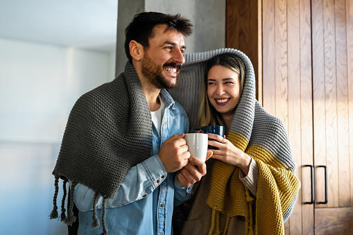 Happy young couple with hot drinks cuddling under warm blanket at home. People happiness concept.