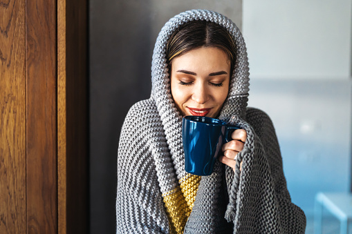 Happy young woman at home in winter wrapped herself in a warm cozy blanket, holding a cup of hot drink