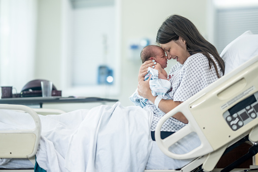 A new Mother sits up in her hospital bed shortly after delivery as she holds her newborn out in front of her and studies his features.  She is wearing a hospital gown and is laying in her hospital bed with the inafnt.