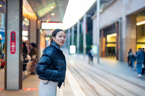 Asian woman waiting for the train at railway station in the city in evening. Attractive girl traveler enjoy urban outdoor lifestyle travel city street by railroad transportation on holiday vacation.