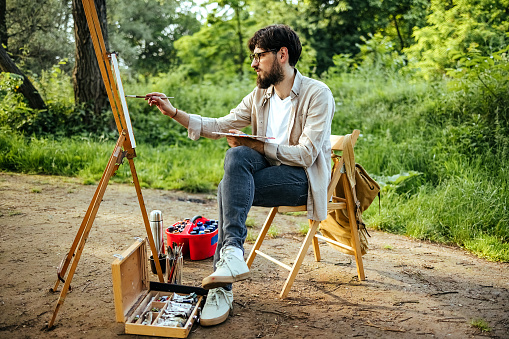 Young male artist painting in nature while sitting on a chair