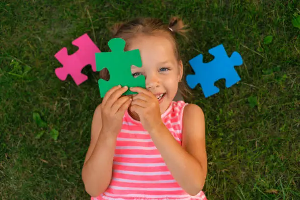 A cute beautiful preschool girl is lying on the grass on the lawn covering her face with puzzle pieces laughing merrily. Preparation for school, development and education