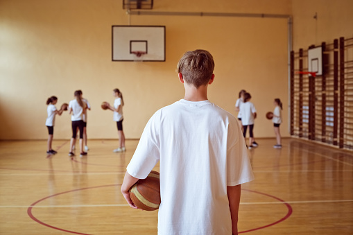 Teenage boy wearing white t-shirts holding basketball balls looking at their defocused friends in the background.