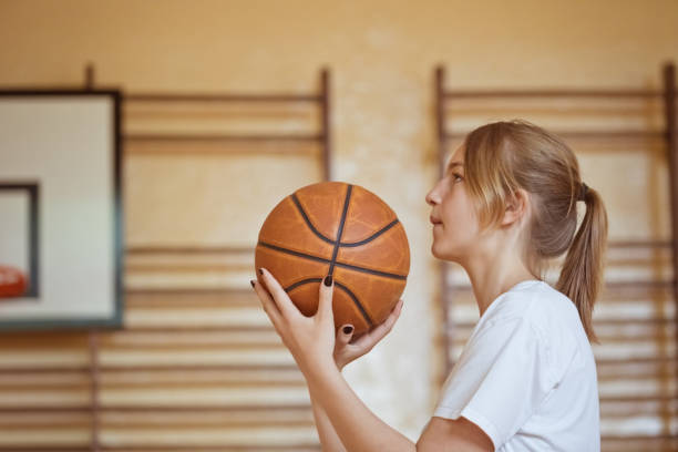 Side view of teenage girl wearing white t-shirts  practicing basketball throws in gym.