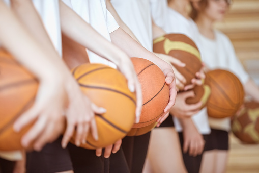 Group of teenager wearing white t-shirts and black shorts standing in row in gym and holding basketball balls. Close up of hands, unrecognizable people.
