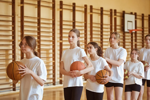 Group of teenage girls wearing white t-shirts and black shorts standing in row in gym and holding basketball balls.