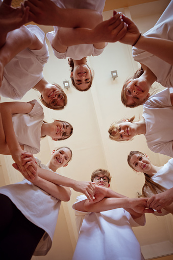 Group of teenage girls and boy wearing white t-shirts standing in circle in gym, holding hands and smiling at camera.