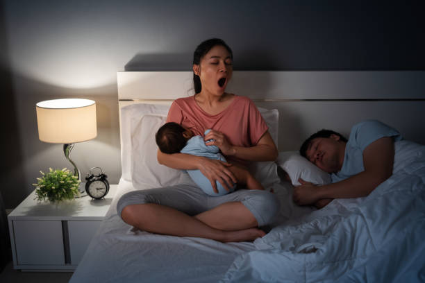 mère fatiguée bâillant et allaitant le bébé newborm pendant que son mari dort sur le lit la nuit - baby yawning asian ethnicity newborn photos et images de collection