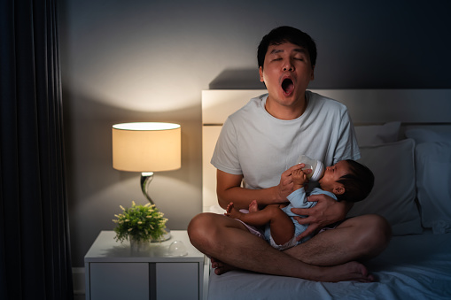 sleepy father yawning and feeding milk bottle to newborn baby on a bed at night