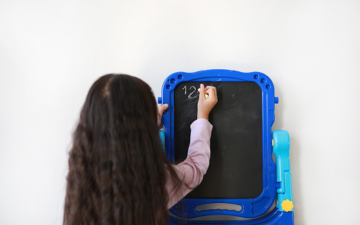 Little Asian child girl writing on blank black board over white background. Education concept. Rear view.
