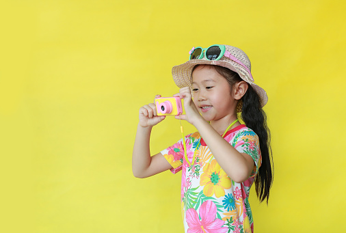 Little girl taking photo by toy camera. Child tourists in floral pattern summer dress and hat with sunglasses isolated over yellow background.