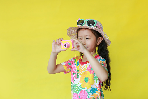 Attractive asian little girl in floral pattern summer dress and hat with sunglasses taking photo by toy camera isolated on yellow background. Fashion of Holiday and summer concept.