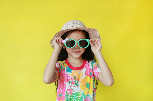 Portrait asian child girl in floral pattern summer dress and hat wearing a sunglasses on yellow background. Fahion of holiday and summer concept