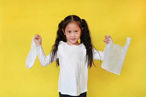 Cute asian little child girl holding a white thin polythene plastic bag isolated on yellow background. Reduce global warming concept.