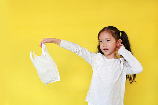 Pretty asian little girl needless a white thin polythene plastic bag for Reduce or zero waste. Kid holding a plastic bag isolated on yellow background. Child and Pollution concept.