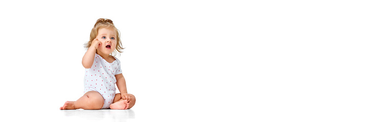 Beautiful, calm, curious baby, little girl sitting on floor and looking against white studio background. Smart child. Concept of childhood, motherhood, care, life, birth. Copy space for ad