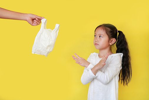 Asian little child girl needless a white thin polythene plastic bag with making X sign her arms for Reduce or zero waste isolated on yellow background. Kid and Pollution concept.