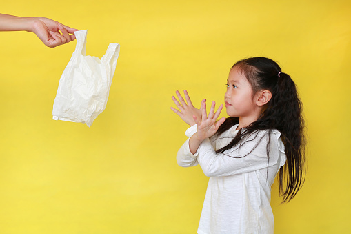Asian little girl making X sign her arms for needless a white thin polythene plastic bag to Reduce or zero waste isolated on yellow background. Pollution concept.