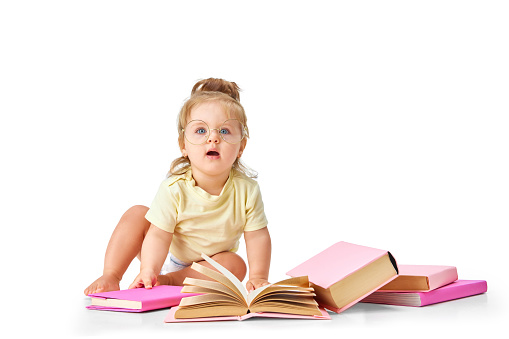 Smart, curious baby. Little girl, toddler sitting on floor with many books around against white studio background. Concept of childhood, motherhood, care, life, birth. Copy space for ad