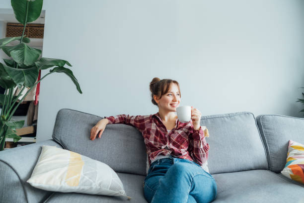 istock Young smiling woman sitting on sofa and looking away while drinking coffee or tea. Young brunette woman relaxing after housekeeping, home cleaning. Portrait of relaxed female resting at home. 1519560470