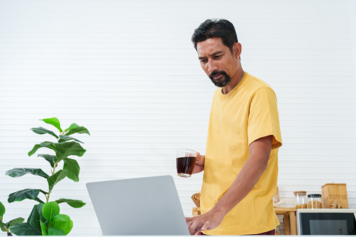 Asian man with beard Standing in the kitchen drinking morning coffee and looking at laptop computers. placed on table Check stock trading on the stock exchange website to get ready for morning trading