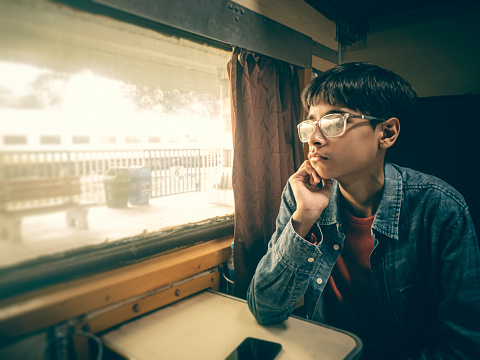 An Asian/Indian boy travels by train in the air-conditioned compartment during his summer vacation. He sits close to the window, looks outside at the view, and contemplates.