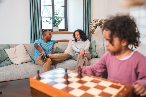 While her father and grandmother chat in the background of their modern apartment, a young multiracial girl happily plays with a chessboard at home.
