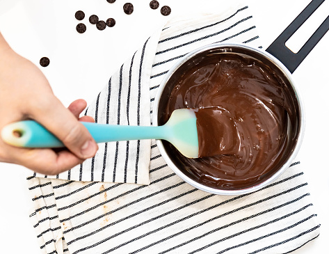 A person's hand uses a spatula to stir to melt chocolate in a saucepan before baking cake and cookies. Pasty and bakery chef mix ingredients in the pot and prepare for homemade dessert. Cooking object on napkin.