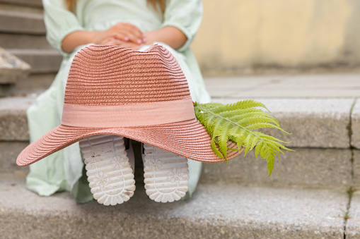 A girl in a romantic dress sits on the stone steps, wearing a pink hat with a fern feather on her legs. High quality photo