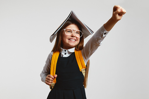Funny young schoolgirl acting like superhero, holding open book on head and raising up fist while pretending flying on white background