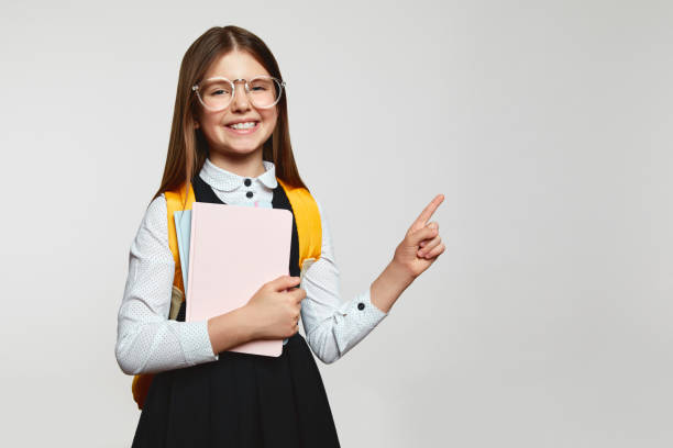 istock Schoolgirl in uniform with yellow backpack holding textbooks while pointing to free empty space 1519918373