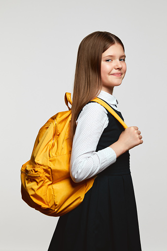 Vertical photo of side view schoolgirl with yellow backpack smiling and looking at camera while against white background. Back to school concept