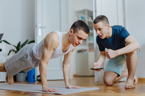 Identical twin brothers doing exercise at home in the morning. One is helping the other by holding his legs while he does sit ups and motivating him.