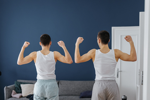 Back view of two teenagers showing their body strength while standing together in the living room.