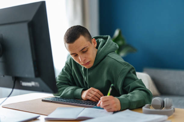 Portrait of a Young Man Learning From Home Portrait of a young male, student, sitting at his desk studying for school or following an online class on his digital tablet. He is taking notes in his notebook. He is doing online class or a course. two men same clothes stock pictures, royalty-free photos & images