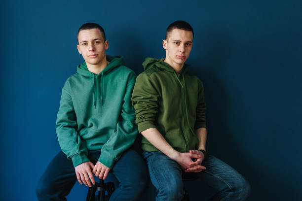 Portrait of Serious Young Twin Brothers Sitting Against the Wall Portrait of two identical twin brother in green hoodies, sitting on the chair against the dark blue wall. They are looking serious facing the camera. and smiling. two men same clothes stock pictures, royalty-free photos & images