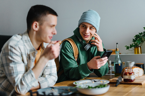 Twin brothers sitting at the kitchen counter and eating their breakfast. One of the brothers is dressed up and ready to go. He has a backpack, bluetooth headphones and a hat. He is looking at his twin, smiling while holding his smart phone.