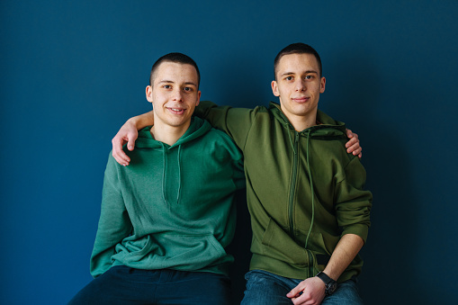 Two smiling teenage boys hugging while sitting on chairs in front of the blue wall and looking at camera.