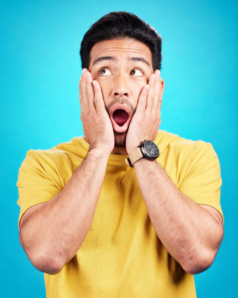 Wow, man and shocked at surprise in studio with announcement, hands on face and mouth open. Male model person on a blue background while scared or thinking of gossip, news or sale with comic emoji wow, hombre y sorprendido por la sorpresa en el estudio con el anuncio, las manos en la cara y la boca abiertas. persona modelo masculina sobre un fondo azul mientras está asustada o pensando en chismes, noticias o ventas con emoji cómico