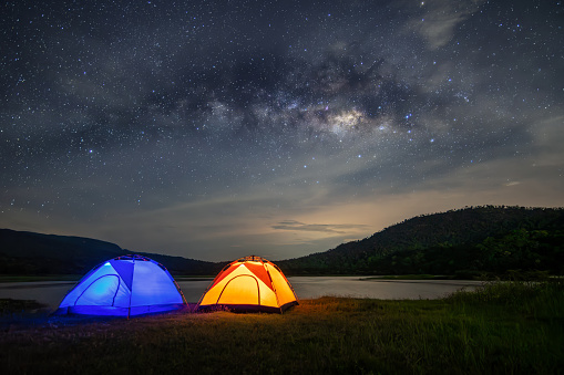 Panorama of the night sky with camping tents and lakes, surrounded by mountains. Milky way and stars on dark background with noise and grain. Long exposure shot with white balance selected.