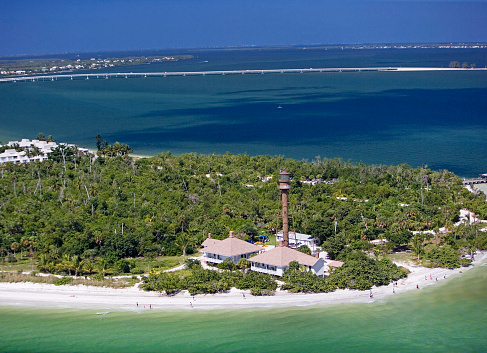 An aerial view of Sanibel Island in southern Florida on a sunny day.  The ocean water is in the foreground, and there are people scattered along the white sandy beach.  There is a great amount of lush vegetation on the island.  A long bridge, the ocean and a blue sky are in the background.