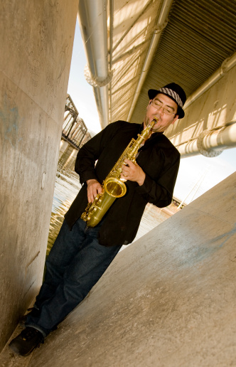 A saxophonist plays outdoors in an urban setting under a bridge.