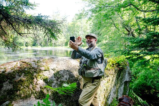 A mature angler outside by a beautiful trout stream using a cell phone.