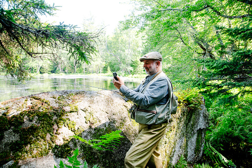 A mature angler outside by a beautiful trout stream using a cell phone.