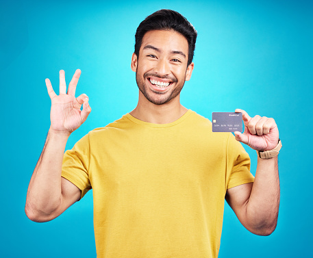 Credit card, okay and portrait of man isolated on blue background for shopping, e commerce and payment loan. Happy asian person, excited for banking solution, financial savings and ok emoji in studio