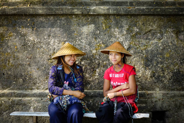 Portrait of three  sitting northern thai women who are wearing traditional clothes from local area. Scene is in rural town Pangkhum in Amphoe Samoeng in eastern Chiang Mai province which is around a 100 years old. Women are smiling. They are wearing purple color based clothes. One is wearing headwear.  In background are examples of traditional loomed thai clothes and scarfs  handmade in this town. Town produces local handmade traditional thai textiles.