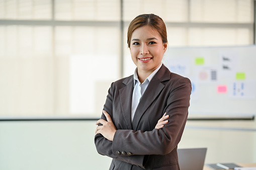 A confident and professional millennial Asian businesswoman or female manager in a formal business suit stands with the meeting room with her arms crossed.