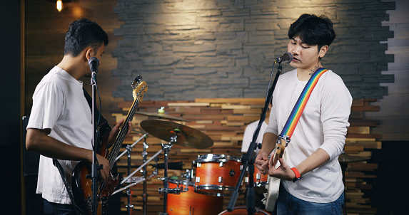 Portrait of Young Asian band of musicians plays during a rehearsal in studio illumination. Bass guitar player, electric guitar player and drummer behind the drum set. Music and entertainment concepts.