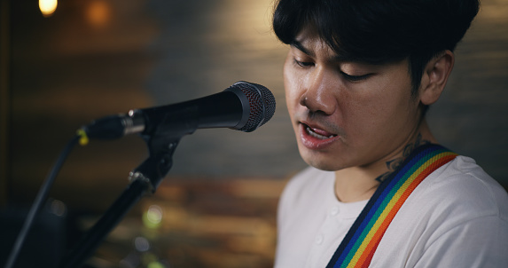 Portrait of Young Asian male musicians in casual clothes playing electric guitar while vocalist singing song into microphone during a rehearsal in studio illumination. Music and entertainment concepts