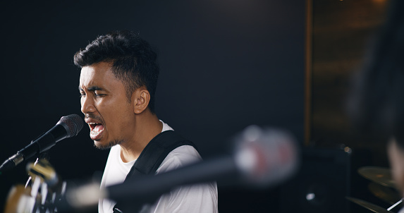 Portrait of Young Asian male musicians in casual clothes playing electric guitar while vocalist singing song into microphone during a rehearsal in studio illumination. Music and entertainment concepts